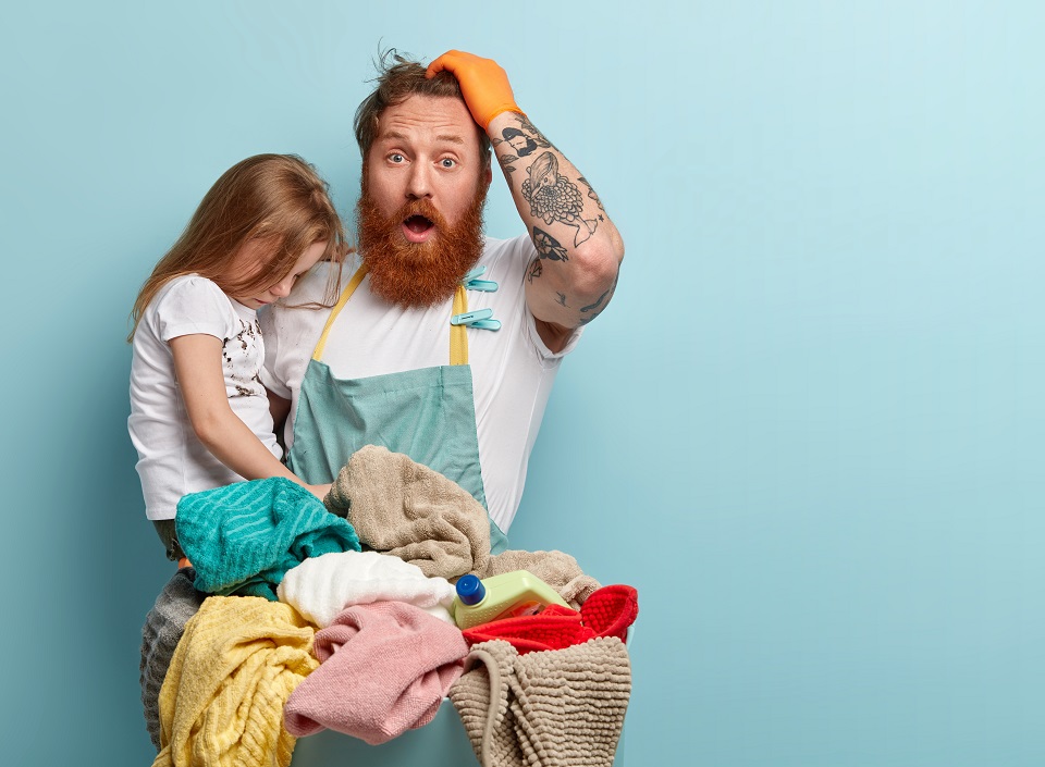Emotional red haired guy keeps hand on head, opens mouth widely, shocked to have much work about house, has to play with child and do household duties, isolated over blue studio wall, blank space